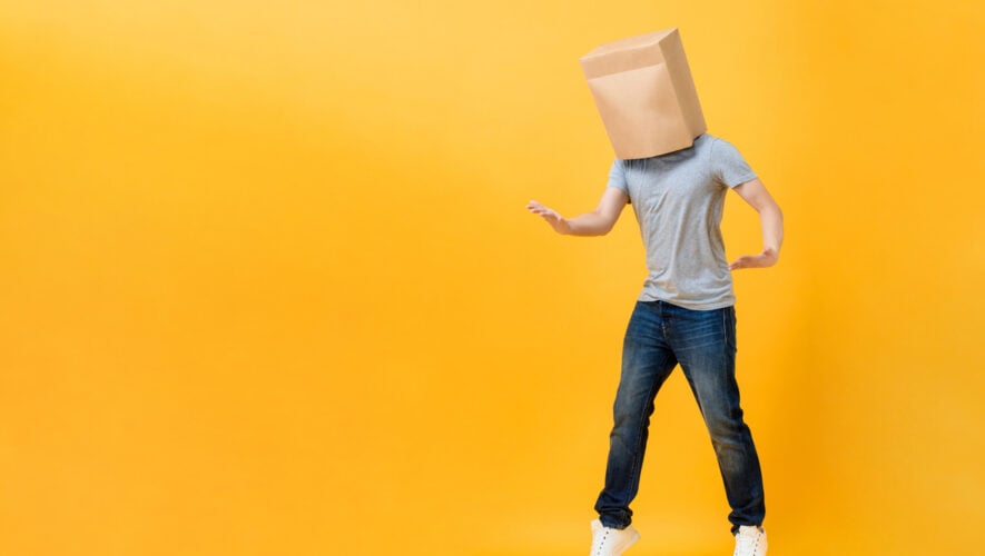 Anonymous man with head covered with paper bag opening hands in yellow studio isolated background